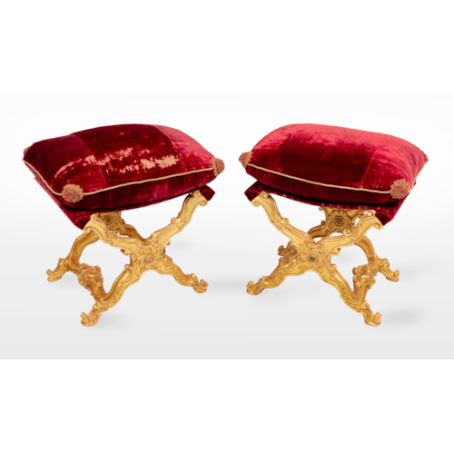 A pair of Italian giltwood carved folding stools, with a rectangular loose cushion seat, in a red silk velvet, above ‘X'-frame baluster supports, carved with scrolls, centred by a patera, joined by shaped stretchers centred by a stylised scallop-shell, on outward scrolled feet.